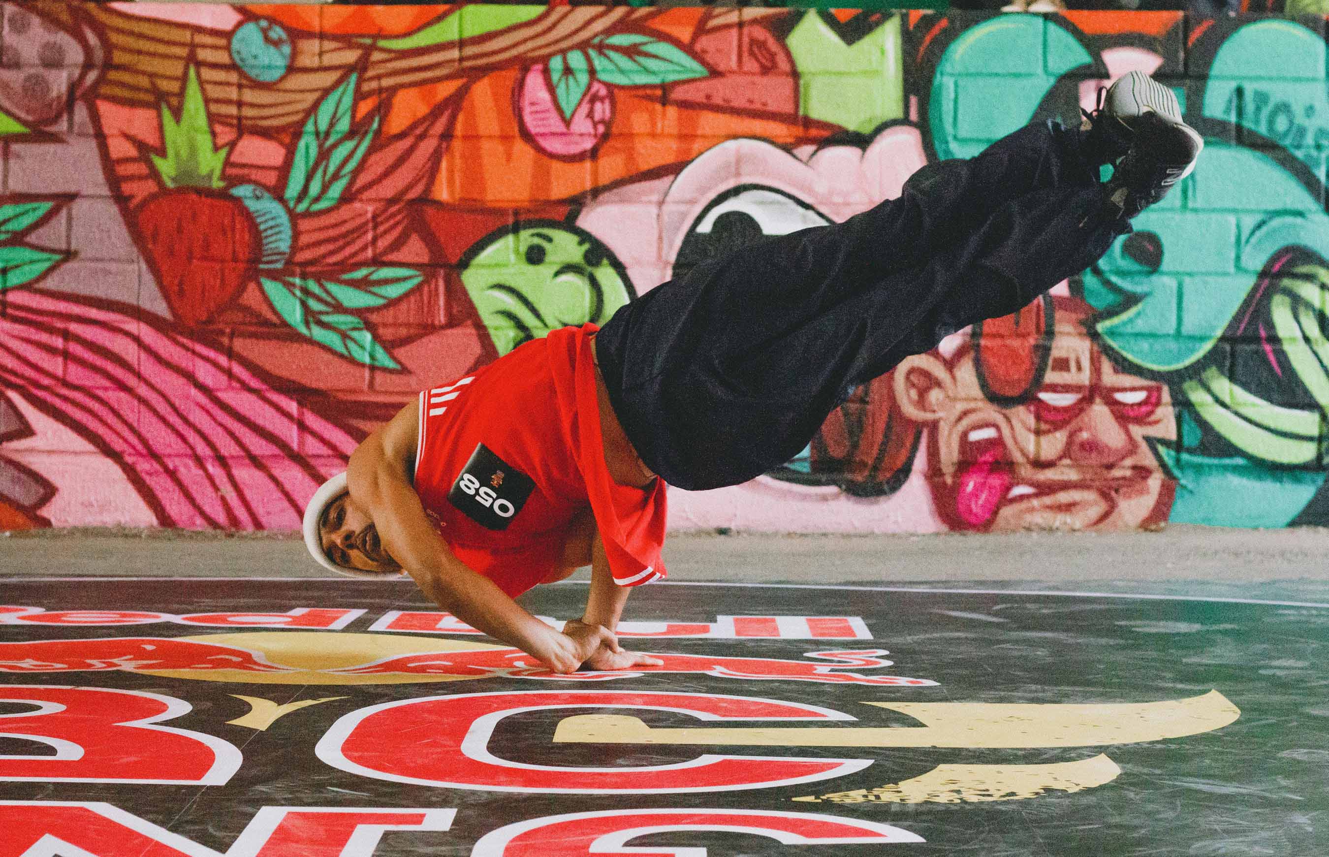 Cours de capoeira, pratiquant en t-shirt rouge réalisant une figure acrobatique en équilibre sur une main devant un mur couvert de graffitis